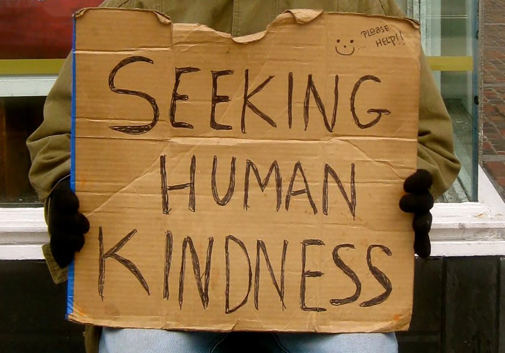 A man holding a cardboard sign that says seeking human kindness.