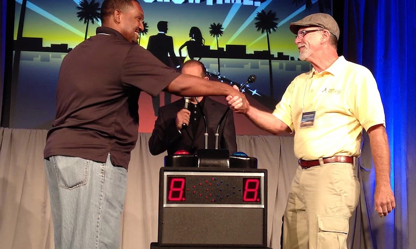 Two men shaking hands in front of a clock to celebrate trivia day.