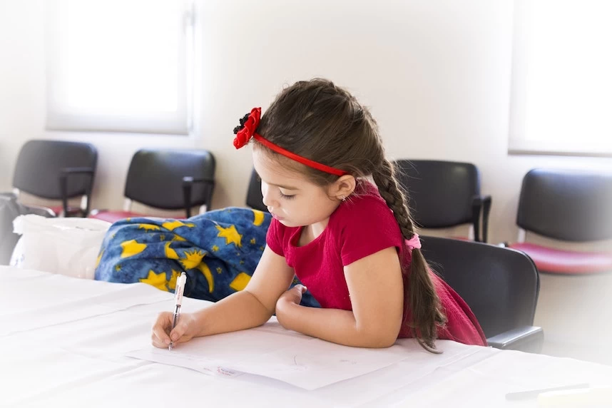 A little girl sitting at a table and writing on a piece of paper.